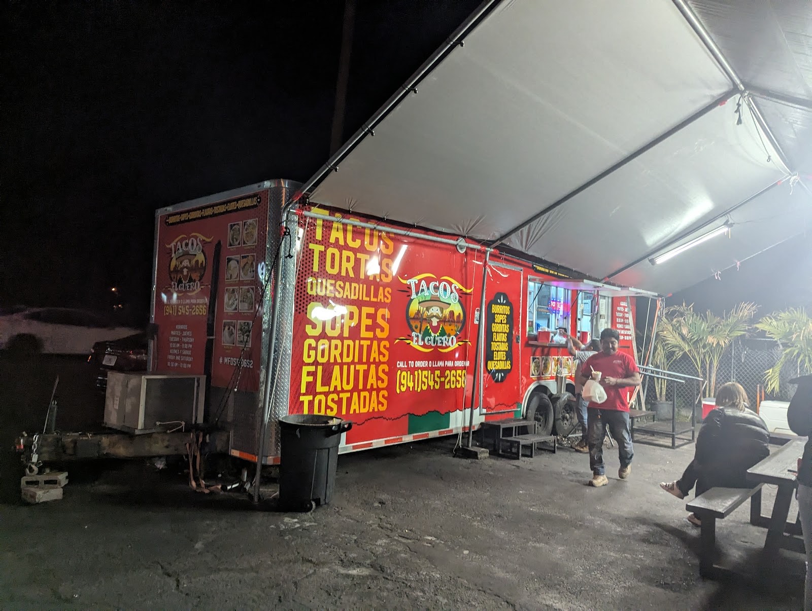 The red Tacos El Güero truck lit up at night under its awning with a guest carrying a takeout bag